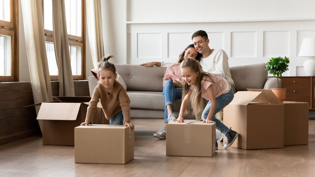 Family with kids moved to first house, while parents resting sitting on comfy sofa two preschool cute daughters play with cardboard boxes pushing it having arranged funny competition. New home concept