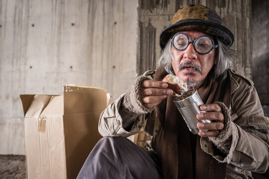 Happy homeless old man sitting eating bread and canned food from donation of kind people. Asian poor beggar senior man wearing glasses, hat, gloves and sweater eating bread because hungry at sidewalk.