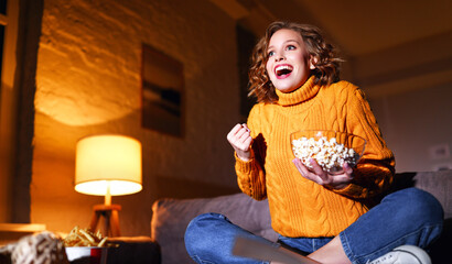 young  delighted cheerful woman with popcorn laughs and watches  movie on  TV   at home in evening ...