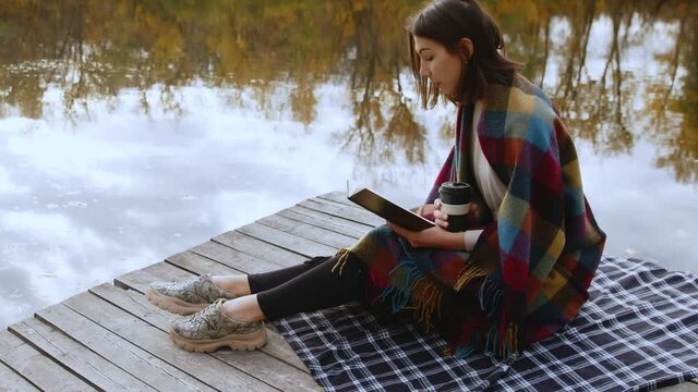 Young beautiful woman wint blanket on shoulders reads book and drinks coffee while stits po piea by the river