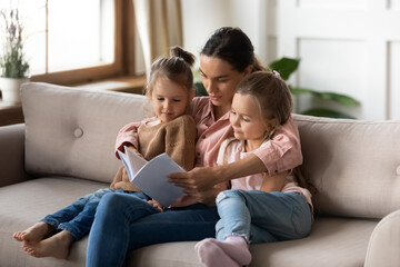 Two cute daughters enjoy interesting fairy tale story read by mommy, sitting together on couch in living room. Hobby and leisure funny time, development and education of children by parent at home