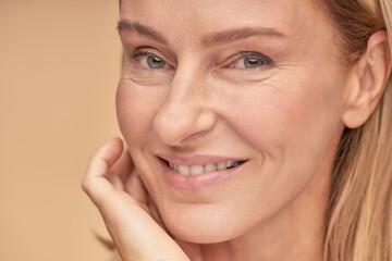 Anti aging skin care. Close up portrait of a beautiful middle aged woman touching her face and smiling at camera while posing over beige background in studio