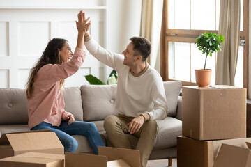 Happy wife and husband sit on couch near heap of belongings in boxes, giving high five congratulate...