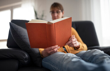 Young brunette woman sitting relaxed in an armchair in a living room reading with picture focus on...