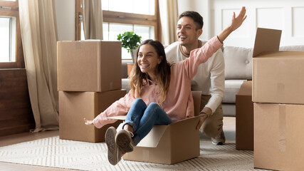 Overjoyed husband pushing playful wife seated inside of cardboard box raised overstretched arms,...