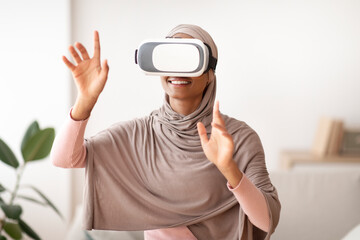 Naklejka premium Black lady in traditional hijab using VR headset to interact with virtual reality at home