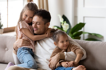 Pleased dad sit on couch with kids, after long separation missed his two little 7s daughters, closing his eyes, hugs them enjoying moment. Family bond and love, Happy Father Day celebration concept