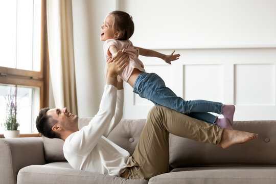 Cute small girl outstretched arms like plane wings imagine like flying in air. Happy dad lies on sofa lifts daughter up on hands play together at home. Active games with kid, travel, vacation concept