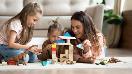 Cute little daughters play with mom family lying on carpet on warm floor in living room, mother help to girls build tower or castle with wooden colorful blocks, free time at home with kids concept