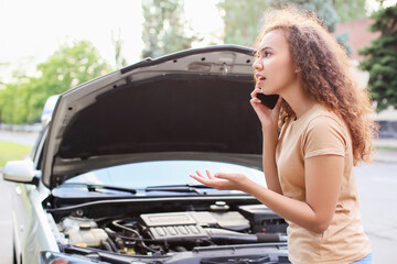 Stressed African-American woman talking by phone near broken car outdoors