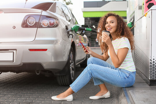 African-American Woman With Mobile Phone Drinking Coffee While Filling Up Car Tank At Gas Station