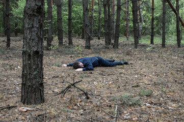 Murder in the woods. The body of a man in a blue shirt and trousers lies on the ground among the trees in the forest. Victim of an attack.
