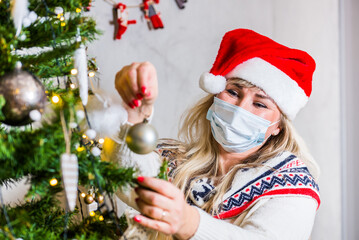 Middle age blond woman with blue eyes in Christmas hat and face protection arranging Christmas tree with decorations