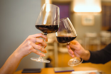 Hands holding a glass of wine clinking. Romance concept of young people celebrating valentine’s day at restaurant toasting with wineglasses. Close up on the hands. Focus on the woman’s hand.