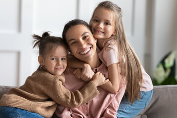 Little 6s 7s beautiful daughters hug young loving mommy, family sitting on couch in living room embracing smiling looking at camera posing for family portrait. Happy Mother Day celebration concept