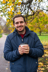 Photo of a young and attractive man enjoying a cup of coffee walking in the park during a day of autumn. 