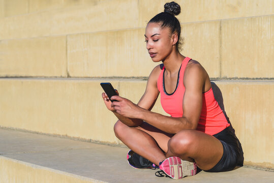 portrait of an attractive african american athlete sitting on concrete steps with crossed legs and using her smart phone - Powered by Adobe