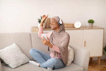 Stay home activities. Pretty Muslim woman in hijab listening to music and dancing on sofa at home, full length portrait