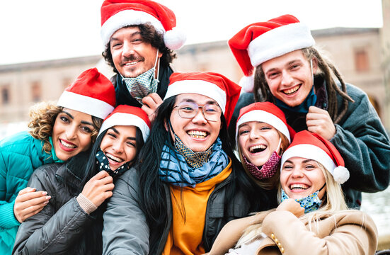 Multiracial people taking selfie wearing face mask and santa hat - New normal Christmas holiday concept with happy friends smiling together outside - Backlight filter with focus on central asian guy