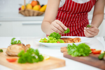 Obraz na płótnie Canvas A beautiful Asian woman places parsley on a salmon steak arranged on a plate with green oak vegetables. Ideas about healthy cooking and weight loss. Soft focus
