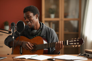 Portrait of talented African-American man singing to microphone and playing guitar while recording...