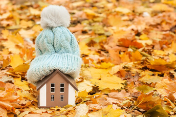 Figure of house and warm hat on autumn leaves outdoors. Concept of heating season