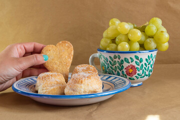 A woman's hand holding a heart shaped cookie surrounded by some traditional Spanish pastries and grapes presented in traditional hand painted ceramic.
