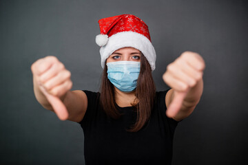Fototapeta na wymiar Girl in black T-shirt, red Christmas cap and medical mask showing thumb down gesture isolated on gray background.