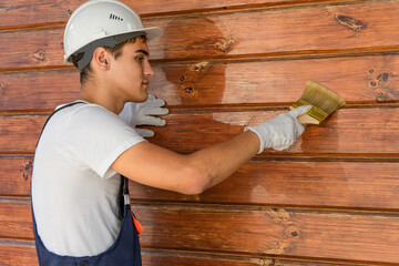 A young painter varnishes the wall of a wooden house . Maintenance of a wooden country house