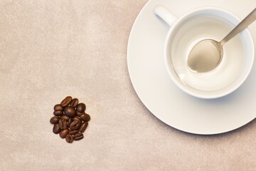 A bunch of roasted coffee beans laying beside a coffee cup with spoon