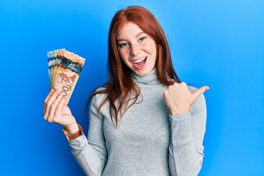 Young red head girl holding canadian dollars pointing thumb up to the side smiling happy with open mouth