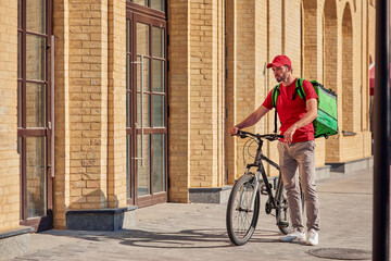 Young male courier in red uniform with thermal backpack standing on the sunny city street