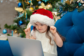 Fototapeta na wymiar A little girl in a hat of Santa Claus grimaces at the camera. Happy New Year on-line on a laptop.