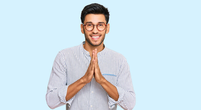 Young handsome man wearing casual clothes and glasses praying with hands together asking for forgiveness smiling confident.