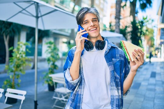 Young hispanic man smiling happy talking on the smartphone and eating sandwich at the city.
