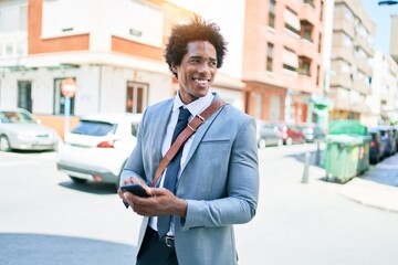 Young african american businessman wearing suit smiling happy. Standing with smile on face using smartphone at town street.