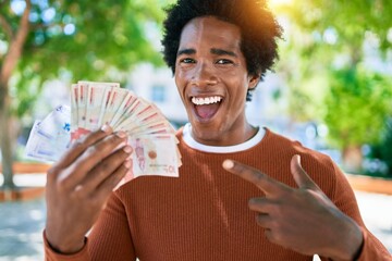 Young african american man smiling happy. Standing with smile on face holding colombian pesos banknotes at town street. - obrazy, fototapety, plakaty