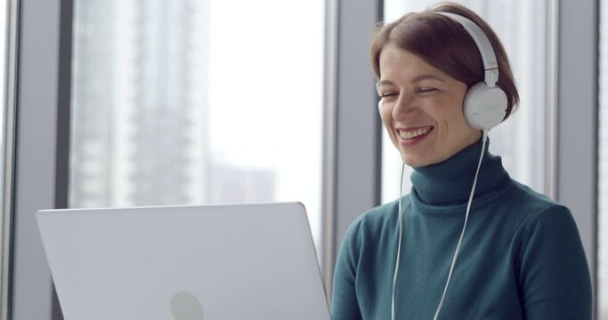Business woman happily talking on a video call using a laptop. Friendly young woman in headphones communicates online video conference remotely. Portrait of a female student at a training webinar.