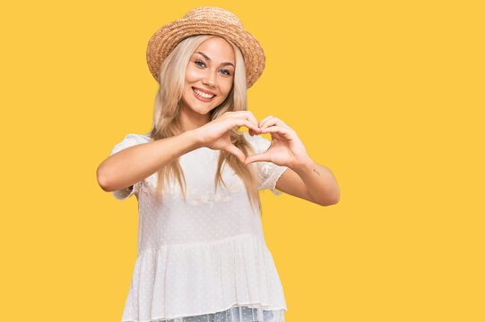 Young blonde girl wearing summer hat smiling in love doing heart symbol shape with hands. romantic concept.