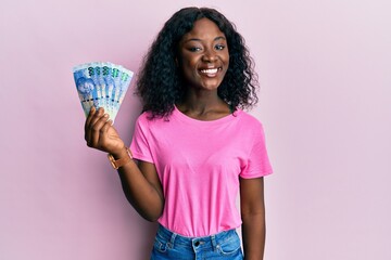 Beautiful black woman holding south african 100 rand banknotes looking positive and happy standing and smiling with a confident smile showing teeth