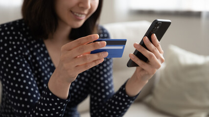 Fototapeta premium Close up smiling Asian woman holding plastic credit card and phone, making secure internet payment, satisfied female customer shopping online, paying, browsing banking service, checking balance