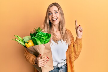 Beautiful caucasian woman holding paper bag with bread and groceries smiling with an idea or question pointing finger with happy face, number one