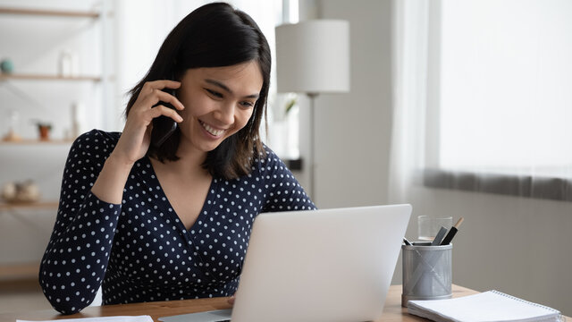 Close up smiling Asian woman talking on phone, using laptop, sitting at work desk, happy businesswoman negotiating project with business partner, friendly manager consulting client, making call