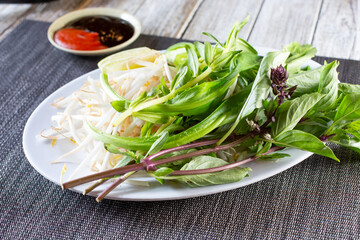 A view of a plate of pho food accessories, featuring Thai basil, mung bean sprouts, lime wedge, hoisin sauce, and sriracha sauce.