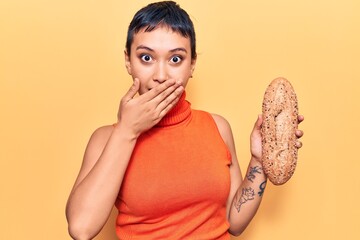 Young woman holding wholemeal bread covering mouth with hand, shocked and afraid for mistake. surprised expression