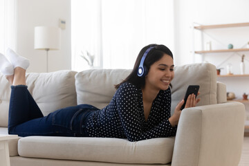 Naklejka premium Smiling relaxed Asian young woman wearing headphones using phone, lying on cozy couch in living room, happy satisfied attractive female chatting or shopping online, listening to music, browsing apps