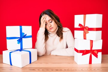 Young beautiful caucasian woman sitting on the table with presents surprised with hand on head for...