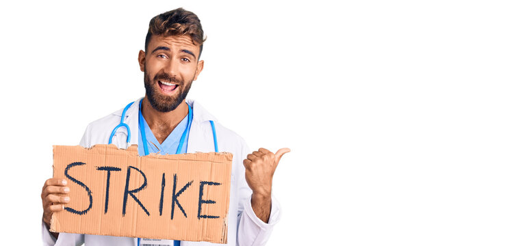 Young hispanic man wearing doctor stethoscope holding strike banner pointing thumb up to the side smiling happy with open mouth