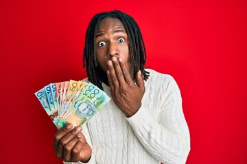 African american man with braids holding australian dollars covering mouth with hand, shocked and afraid for mistake. surprised expression