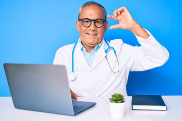 Senior handsome man with gray hair wearing doctor uniform working using computer laptop smiling and confident gesturing with hand doing small size sign with fingers looking and the camera. measure.